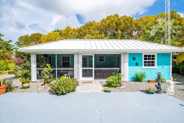 A small, single-story house with a white roof and turquoise walls. It features a front porch with potted plants and a statue.