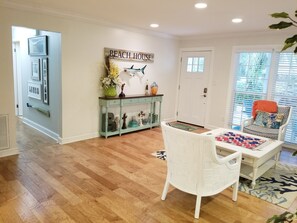 Una habitación con piso de madera, una silla blanca, una mesa con colcha, un tocador con objetos decorativos y una pared con un cartel que dice 'Beach House'.