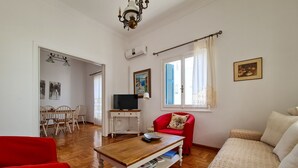 A living room with a wooden coffee table, red armchair, and beige sofa. There is a TV on a white stand, a painting on the wall, and a window with curtains. - My Home in Naxos Town With Sea View (Naxos)