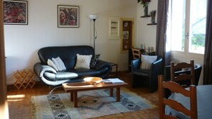 A living room with a black leather sofa, a wooden coffee table, and a dining table with chairs. There is a patterned rug on the floor, a standing lamp, and framed pictures on the wall. - Air-conditioned & heated cottage in picturesque Pernes-les-Fontaines (Pernes-les-Fontaines)