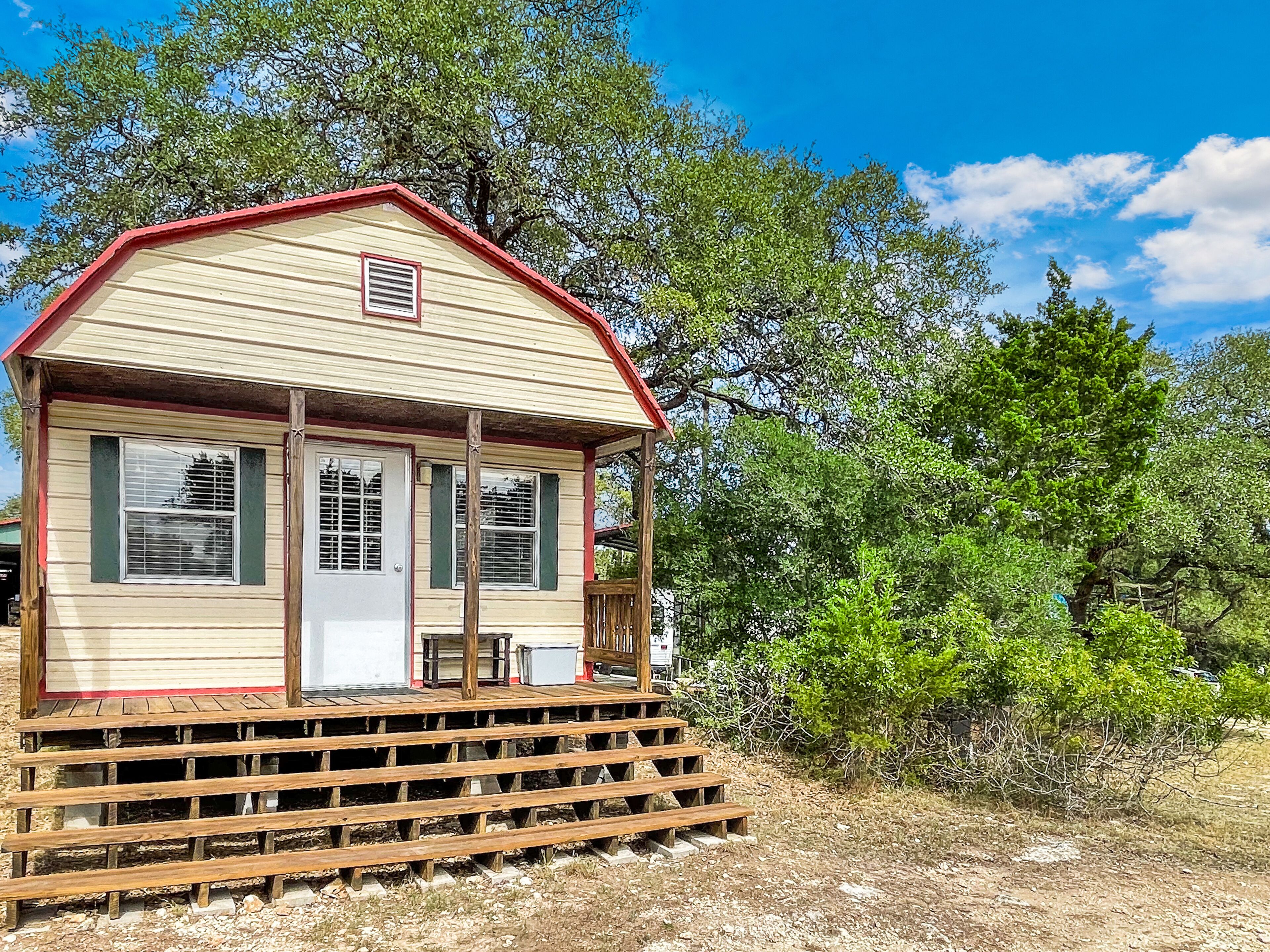 A small, single-story cabin with a red roof and beige siding, featuring a front porch and a small deck.