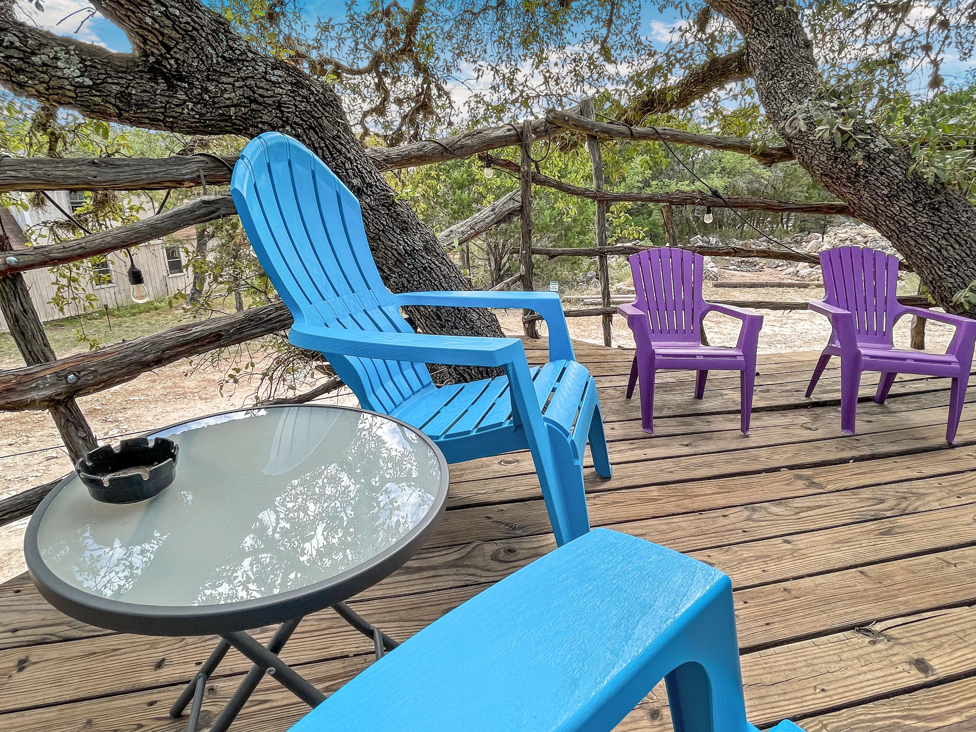 A wooden deck with blue and purple Adirondack chairs, a glass table, and a rustic wooden fence.