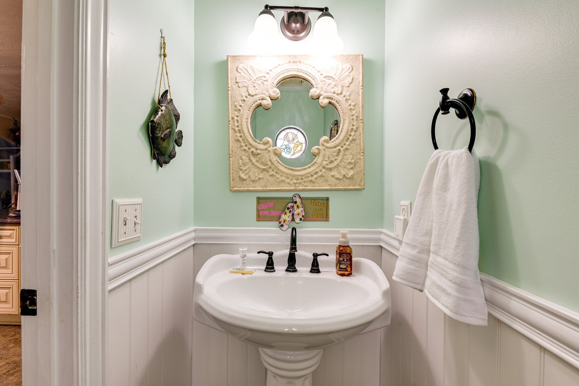 Ein Badezimmer mit einem weißen Waschbecken, einem Spiegel mit aufwendigem Rahmen, einem Handtuchhalter und einem dekorativen Fisch an der Wand.