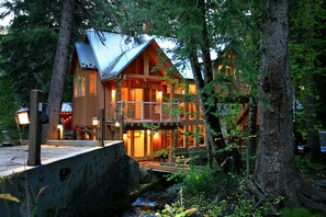A wooden cabin with a balcony, surrounded by trees and a small stream.