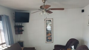 Una sala de estar con un sofá marrón, un sillón reclinable, un tapete estampado, un ventilador de techo y un televisor montado.