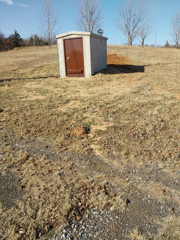 Una pequeña construcción de una sola habitación con una puerta marrón, ubicada en un campo cubierto de hierba con algunos árboles al fondo.
