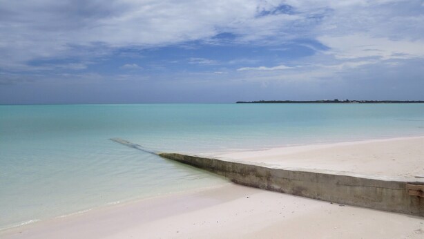 Een strand met helder turquoise water en een betonnen looppad.