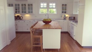 Een keuken met witte kastjes, een houten eettafel, een fruitschaal, een spoelbak, een kookplaat en een raam.