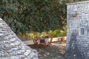 A stone building with a window, a stone wall, and a table with chairs in front of a dense green hedge. - Authentic And Lovely Trulli In Monopoli’s Countryside (Monopoli)