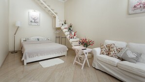 Um quarto com uma cama, um sofá branco, uma mesinha, uma escada e uma prateleira fixada na parede com objetos decorativos.