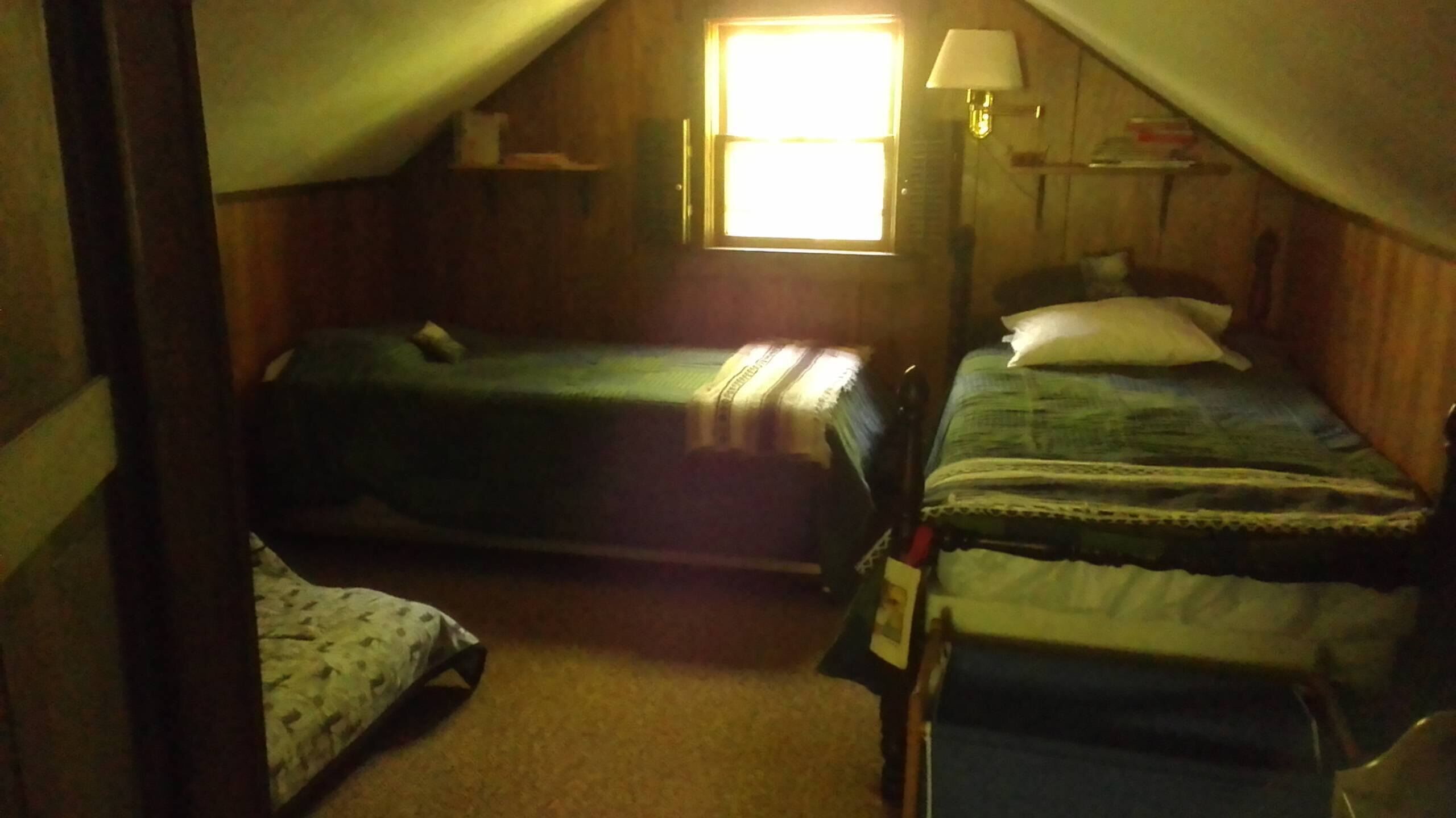 Une petite chambre mansardée, peu éclairée, avec deux lits simples, une fenêtre avec une lampe et une porte en bois.