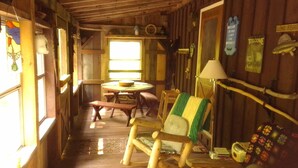 Interior rústico de una cabaña con una mesa de madera, bancos, una silla con una manta verde y amarilla, una lámpara y un cartel de bienvenida.