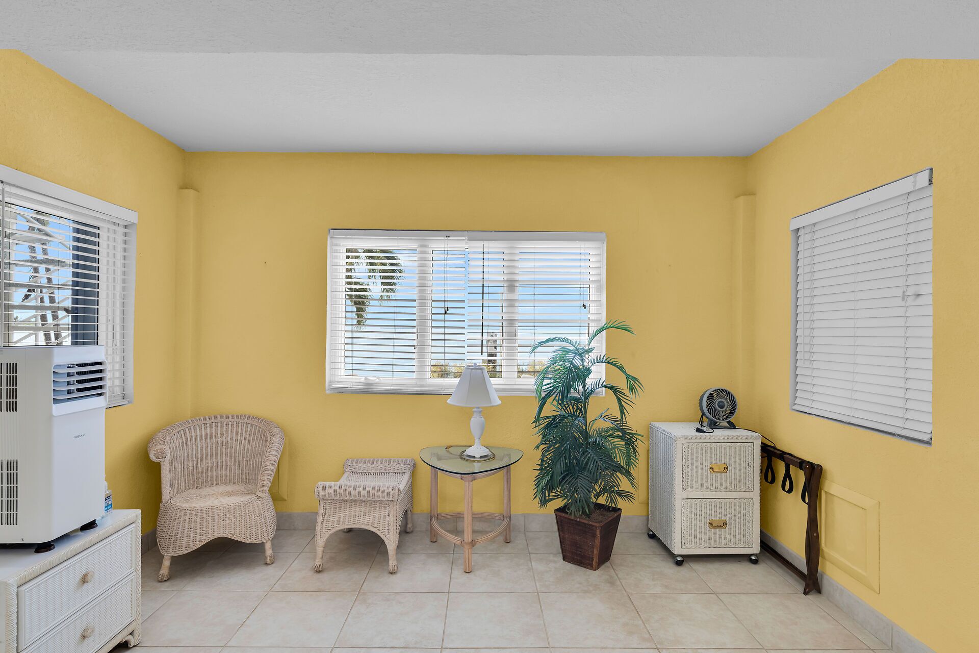 A room with yellow walls, a white ceiling, a wicker chair, a small table with a lamp, a potted plant, a white cabinet, and a window with blinds.
