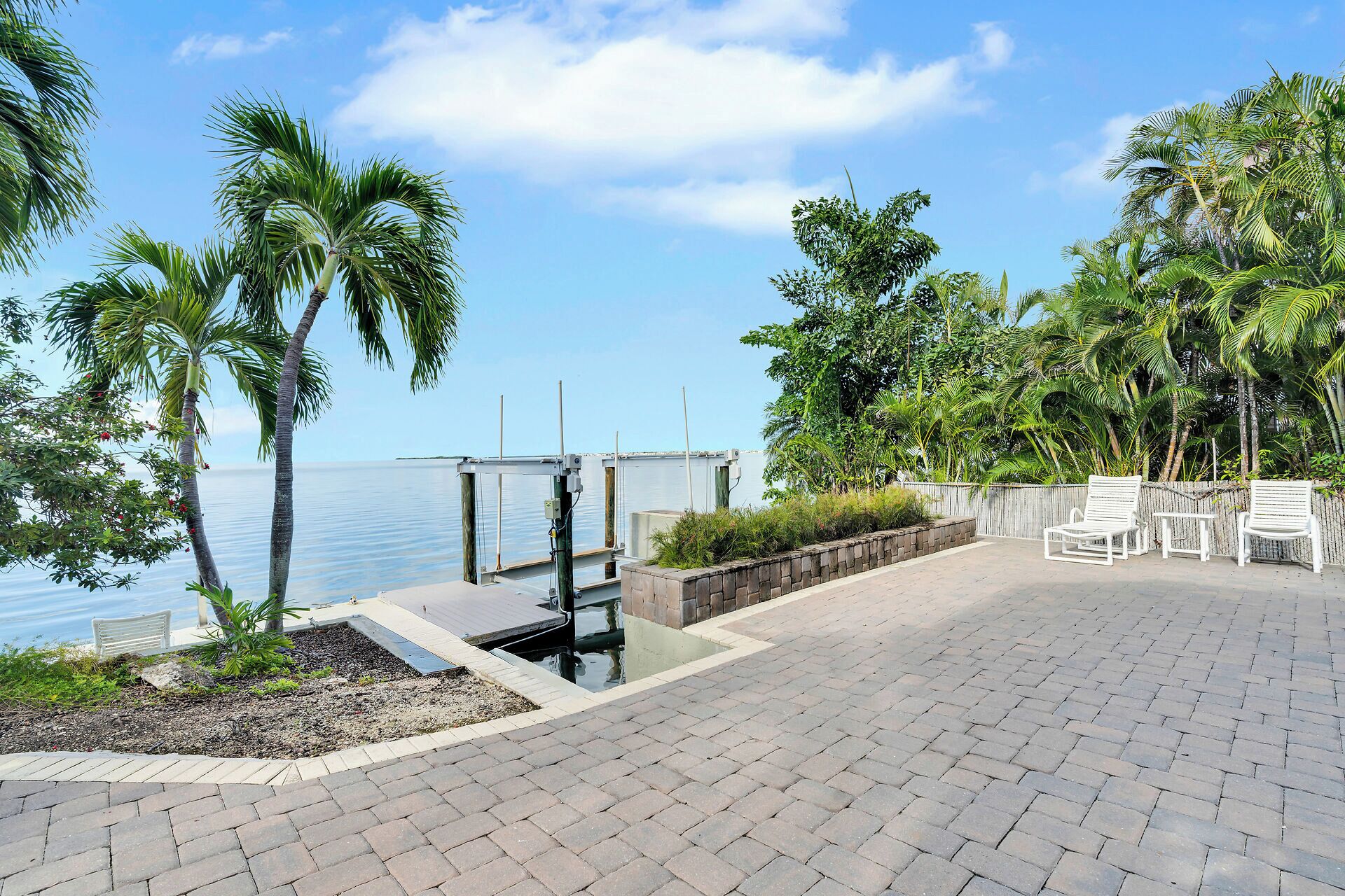 A paved patio with white chairs, a dock extending into calm water, and lush tropical vegetation.