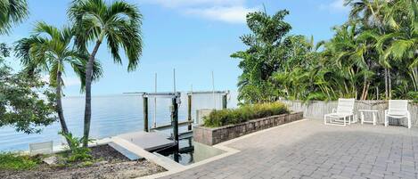 A paved patio with white chairs, a dock extending into calm water, and lush tropical vegetation.