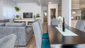 A modern living room with a grey sofa, a dining table with chairs, a flat-screen TV, and a kitchen area with wooden cabinets. - Entire house just for you! (Pula)