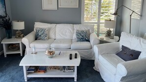 Una sala de estar con un sofá blanco, una mesa de centro y un sillón blanco. Se observan cuadros en la pared, una ventana con persianas y una lámpara sobre una mesita.