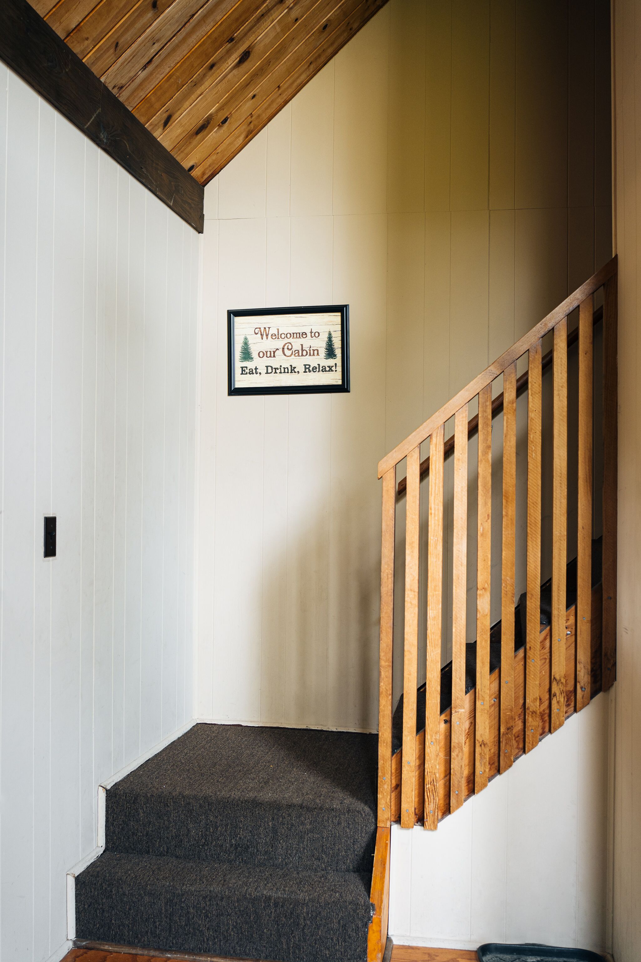 Un escalier avec une rampe en bois et des marches recouvertes de moquette mène à un palier. Un panneau d’accueil encadré est accroché au mur.