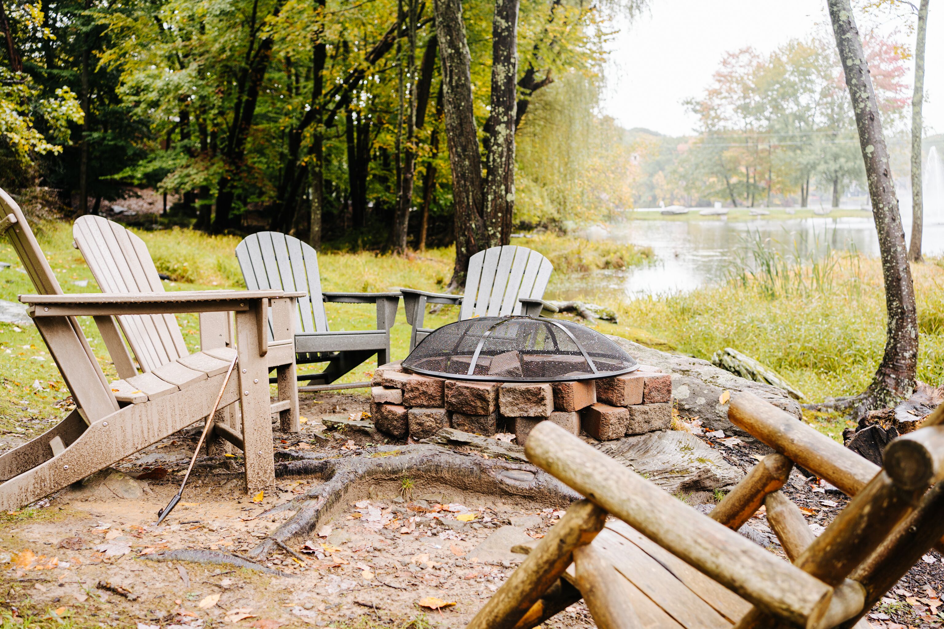 Een rustieke buitenruimte met Adirondack-stoelen en een vuurkorf van steen, omgeven door bomen en een meer.