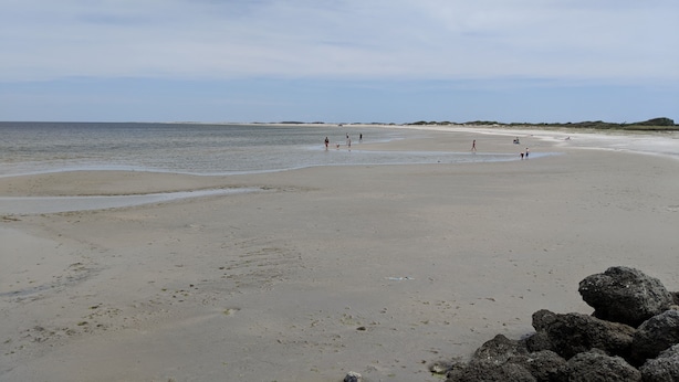 A wide sandy beach with calm water, a few people walking, and rocky outcrops in the foreground. - 2 Bedroom, second row beach cottage .  (Cedar Island)
