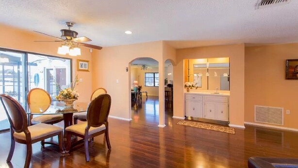 A spacious living room with hardwood flooring, a dining area with a glass table and chairs, a kitchen with white cabinets and a large mirror, and a ceiling fan. - Waterfront Home With Pool (Port Richey)