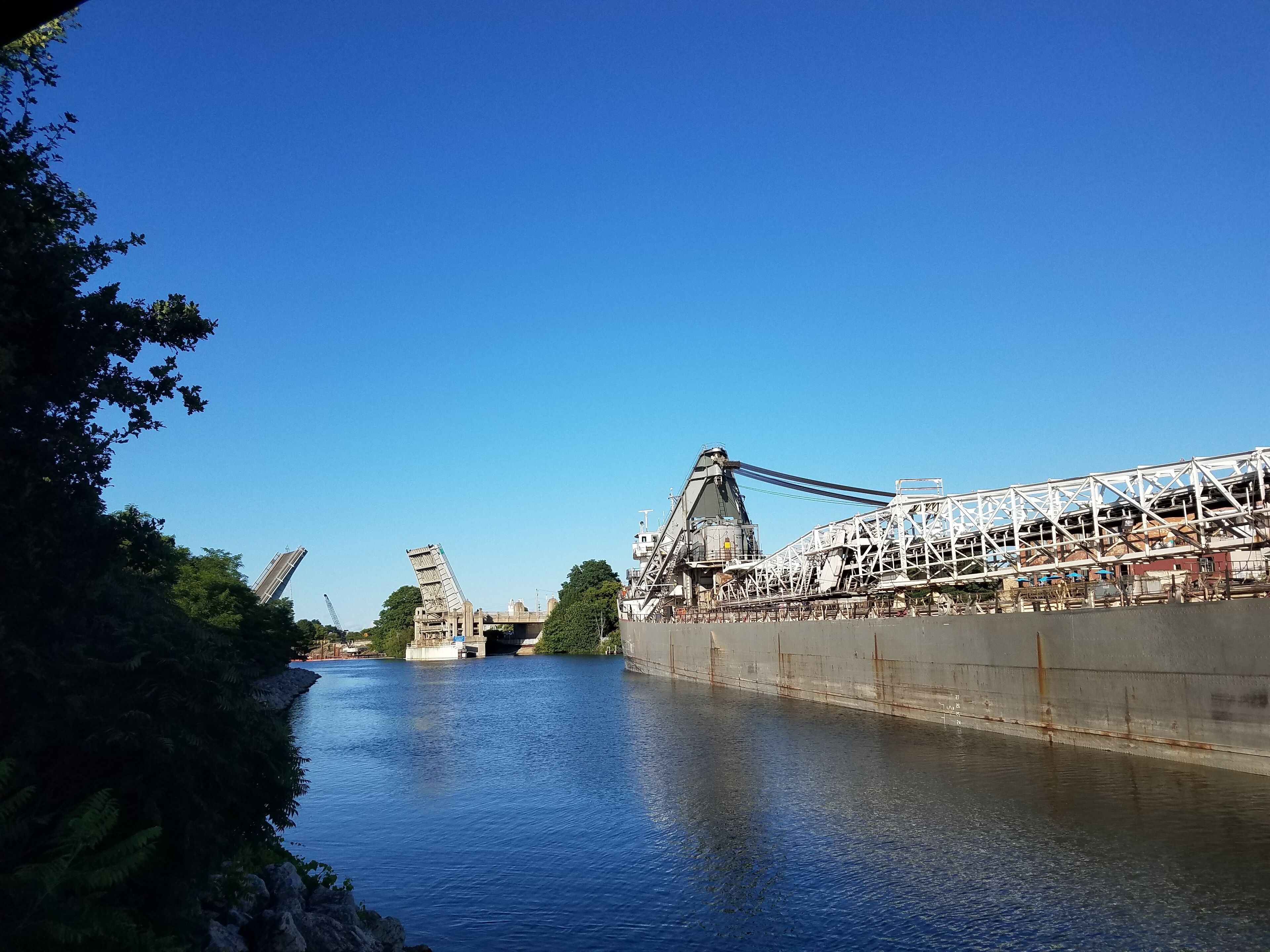 Un pont avec une grande structure industrielle sur la droite, une rivière au premier plan et des arbres sur la gauche.
