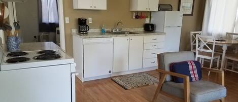 Uma cozinha compacta com armários brancos, fogão e pia. Há uma área de jantar com mesa e cadeiras, e um tapete estampado no chão.