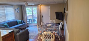 Ein Wohnzimmer mit einer Couch, einem Esstisch mit Stühlen, einem an der Wand befestigten Fernseher und einer Glasschiebetür, die zu einer Außenpatio führt.