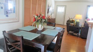 Un comedor con una mesa de madera, sillas y un florero. Se ve una puerta y una lámpara al fondo.