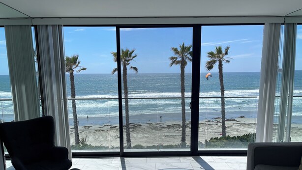 Un salon moderne doté d’une grande porte vitrée donnant sur un balcon qui offre une vue sur une plage bordée de palmiers et sur l’océan.