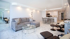 Un salon moderne avec un canapé d’angle gris, une table basse en verre et un pouf en cuir. On y trouve également un coin repas avec une table et des chaises, et une cuisine en arrière-plan.