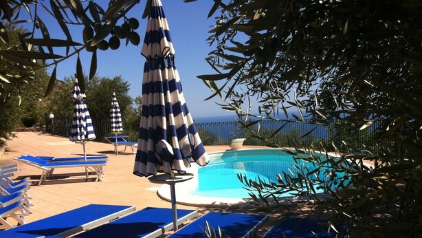 Zona junto a la piscina con sillones azules y sombrillas a rayas, rodeada de olivos y una vista despejada del mar.