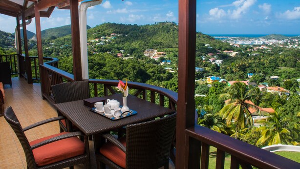 Ein Balkon mit Korbmöbeln, einem Tisch mit einer Teeservice und einer Vase, und Blick auf eine grüne Landschaft mit Gebäuden.