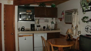 A compact kitchen with a microwave, sink, and small refrigerator. There is a wooden dining table with chairs and a television.