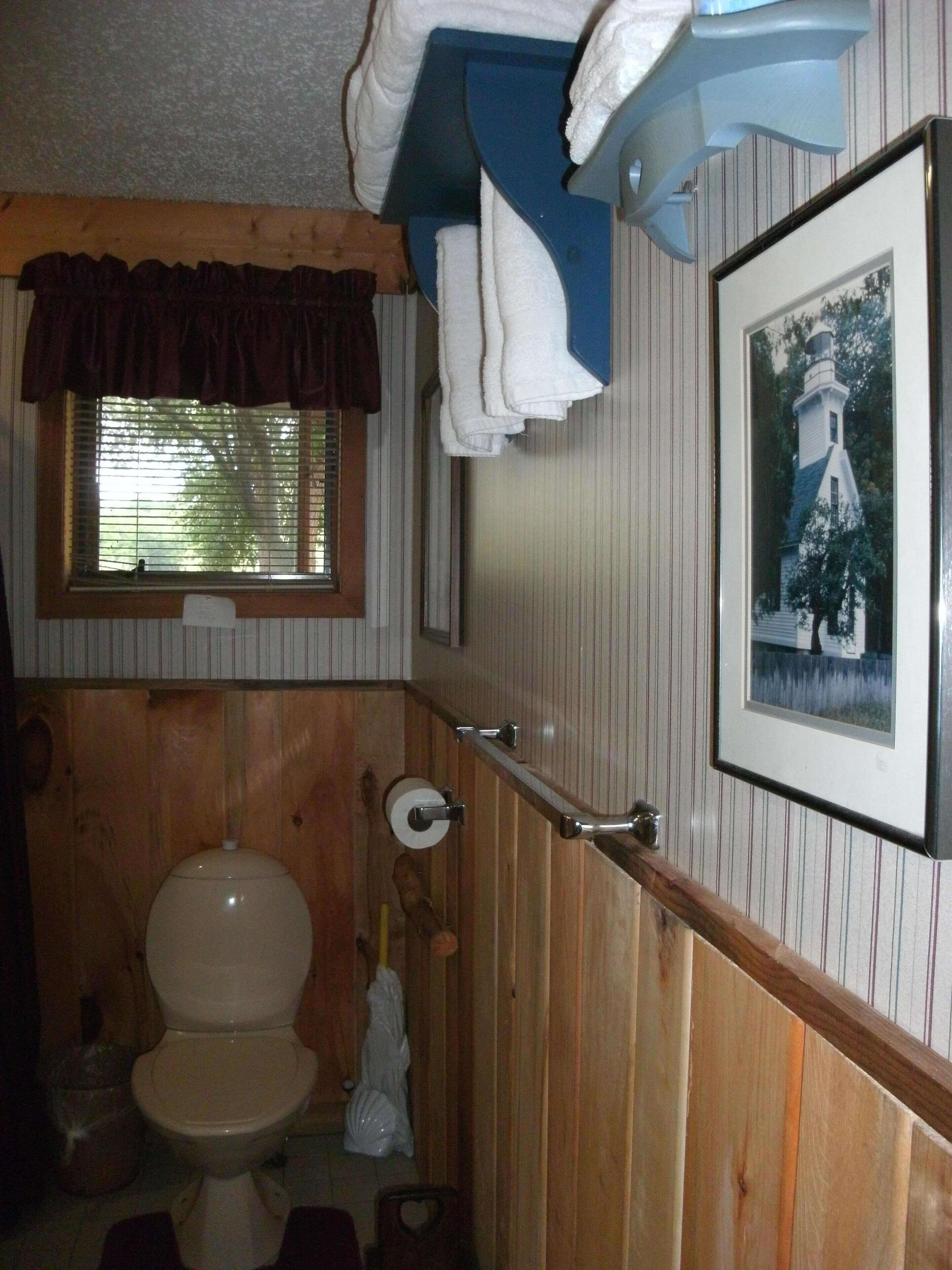 A bathroom with wooden paneling, a toilet, a window with blinds, a towel rack, and a framed picture on the wall.
