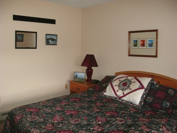 A bedroom with a floral bedspread, a wooden headboard, a nightstand with a lamp, and framed pictures on the wall.