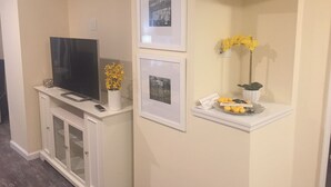 A living room with a flat-screen TV on a white cabinet, a framed picture above the cabinet, and a small shelf with a vase of yellow flowers and a tray of oranges. - Almost Beachfront, 7 houses to the Beach (Ventnor City)
