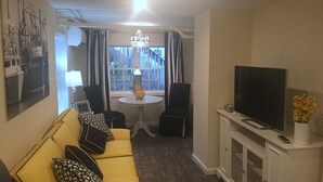 A living room with a yellow sofa, a TV on a cabinet, a chandelier, and a window with curtains. - Almost Beachfront, 7 houses to the Beach (Ventnor City)