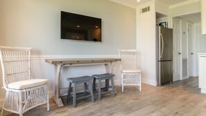Une cuisine avec une table en bois, deux chaises en osier blanches, un réfrigérateur en acier inoxydable et un téléviseur à écran plat fixé au mur.