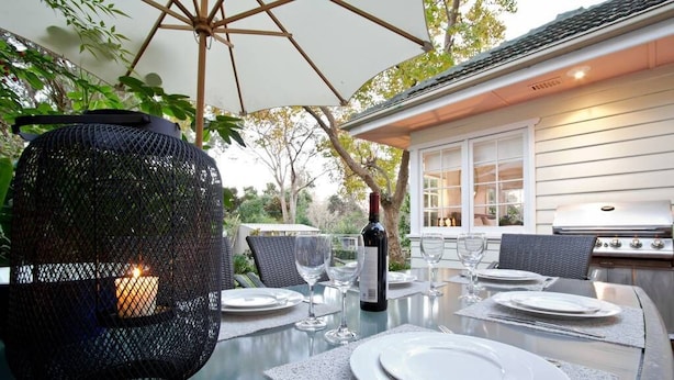 Un área de comedor al aire libre con una mesa dispuesta para cuatro personas, rodeada de sillas de mimbre, una vela en un farol negro y una parrilla al fondo.