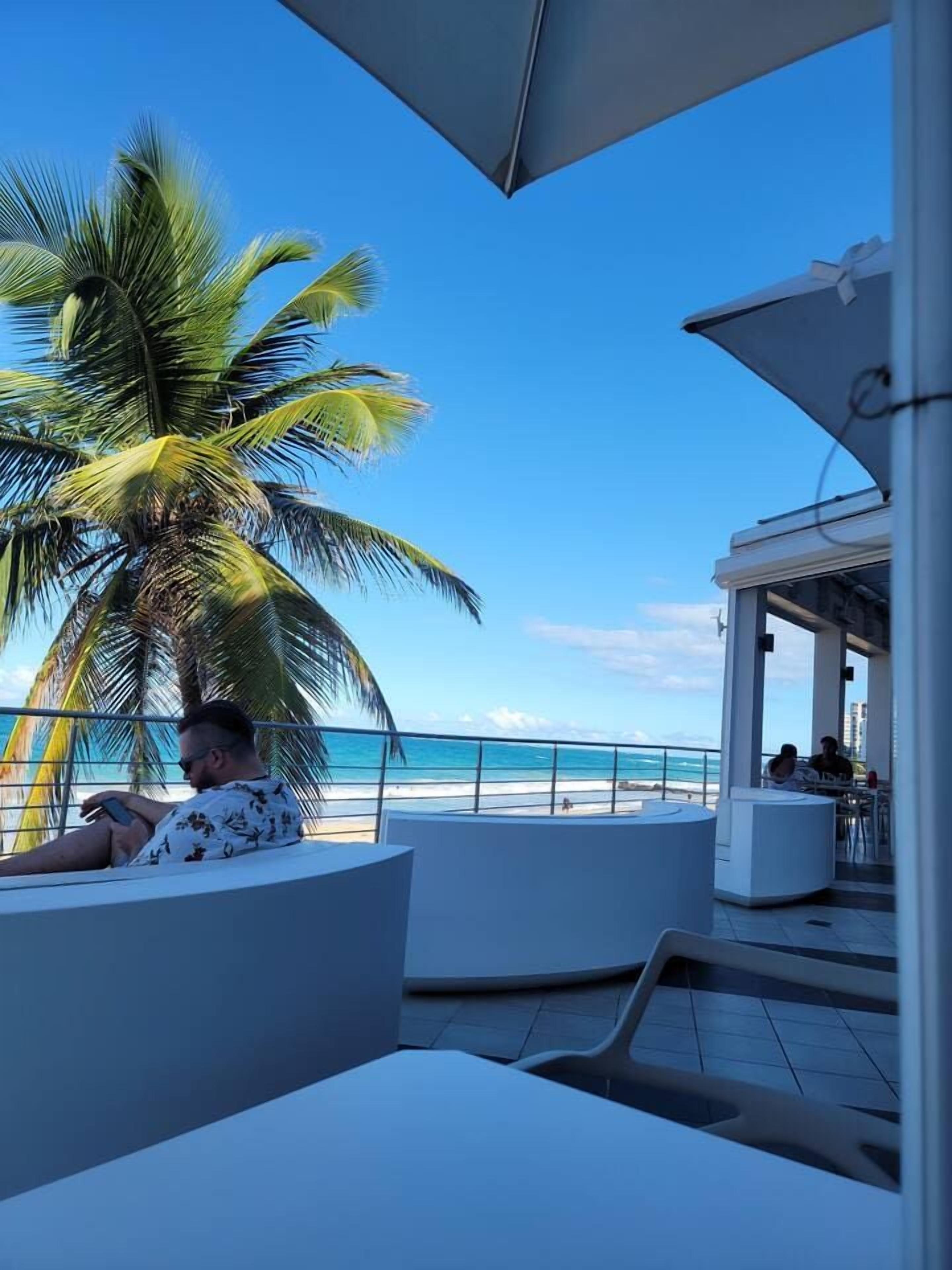 A man relaxing on a white lounge chair with a view of the ocean and a palm tree.