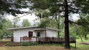 A small white cabin with a porch, surrounded by trees and a grassy area. - Herons Nest at Viewpoint Beach channel on Magician Lake, Sister Lakes, MI 49047 (Sister Lakes)