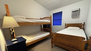 A room with two bunk beds, a desk with a lamp, a window with blinds, and a wall-mounted air conditioner. - Herons Nest at Viewpoint Beach channel on Magician Lake, Sister Lakes, MI 49047 (Sister Lakes)