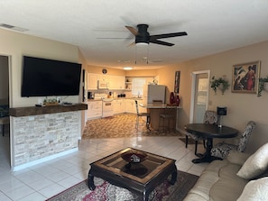 A living room with a flat-screen TV, a coffee table, and a sofa. There is a kitchen in the background with white cabinets and a dining area with a table and chairs.