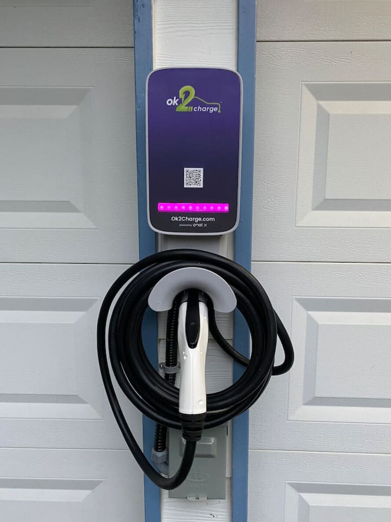 Una estación de carga para vehículos eléctricos con una unidad de carga morada y blanca, un código QR y un cable de carga enrollado.