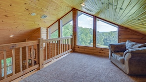 Un chalet confortable avec un intérieur en bois, un balcon doté d’une balustrade et une grande fenêtre donnant sur la forêt.
