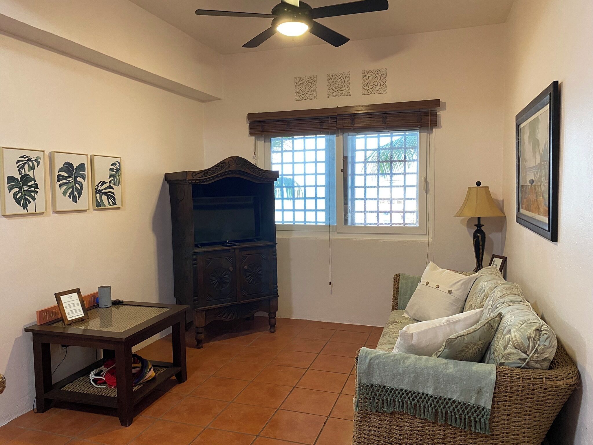 Una sala de estar con un ventilador de techo, un mueble de madera, un sofá, una mesa y una ventana con persianas.