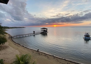 Uma praia com um cais se estendendo sobre águas calmas, um barco atracado à direita e um pôr do sol ao fundo.