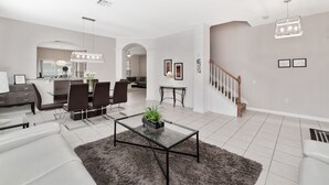 A spacious living room with a glass coffee table, a gray rug, a dining area with chairs, a kitchen in the background, and a staircase.