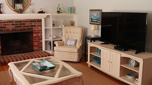 Ein Wohnzimmer mit Kamin, Fernseher, einem Glastisch, einem beigen Sessel und einem Bücherregal.